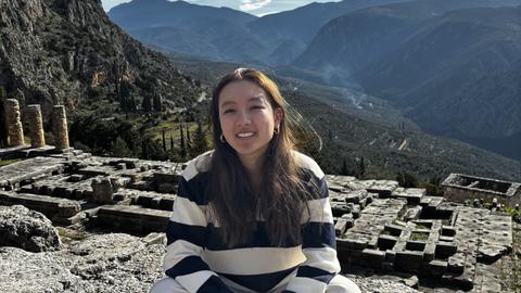 A woman sits on ancient stone ruins with mountains and valleys in the background.