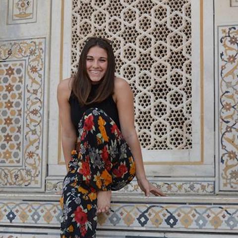 A person smiles while sitting against an ornately decorated wall with intricate geometric and floral patterns.