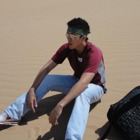 A person wearing sunglasses and a bandana sits on sand dunes with a backpack beside them under the bright sun.