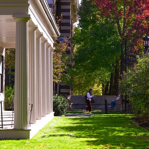 Students walk and relax near a columned building surrounded by trees with autumn foliage.