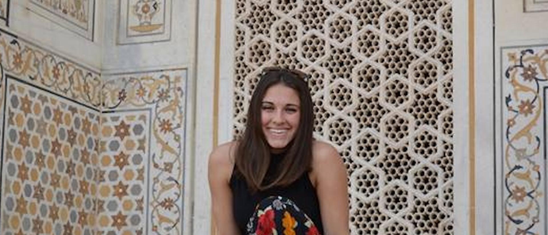 A person smiles while sitting against an ornately decorated wall with intricate geometric and floral patterns.