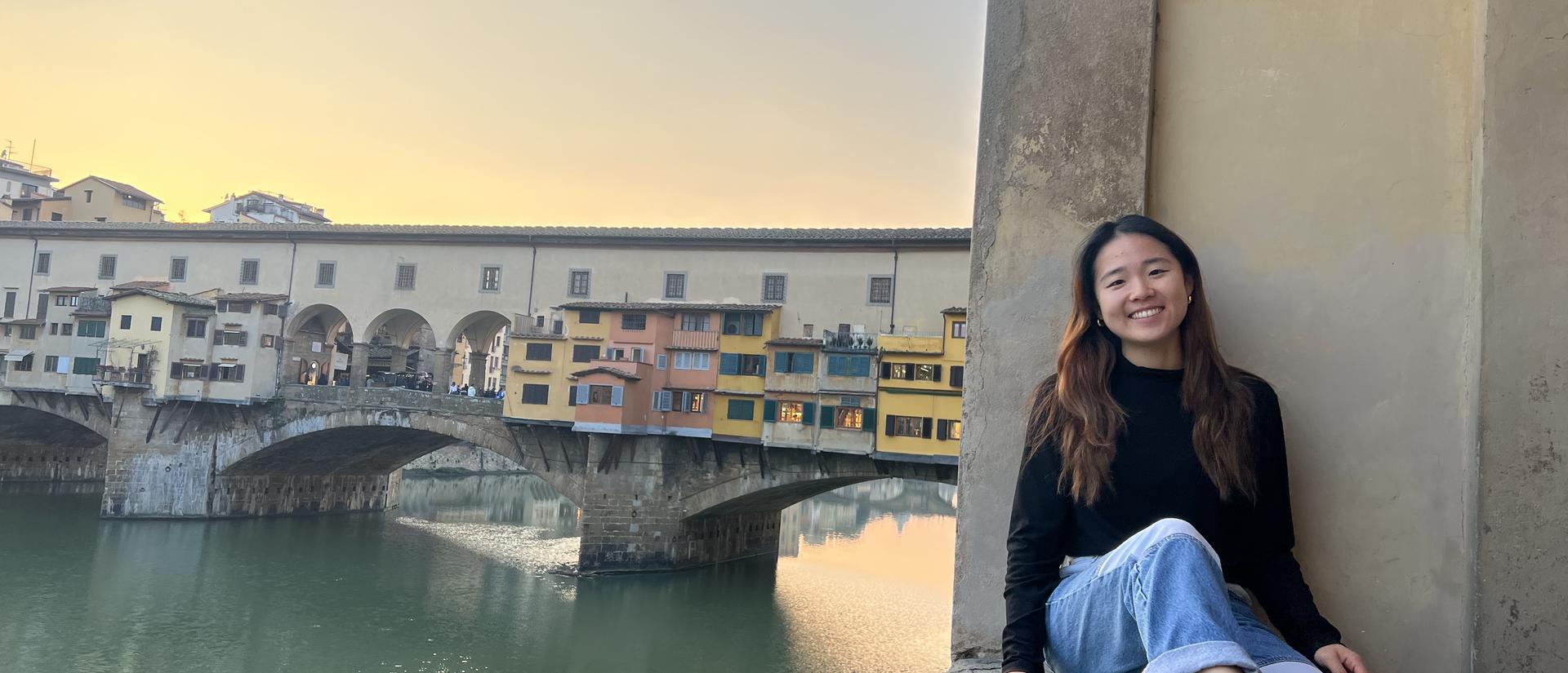 A smiling person sits on a stone ledge with the historic Ponte Vecchio bridge in Florence, Italy, in the background at sunset.