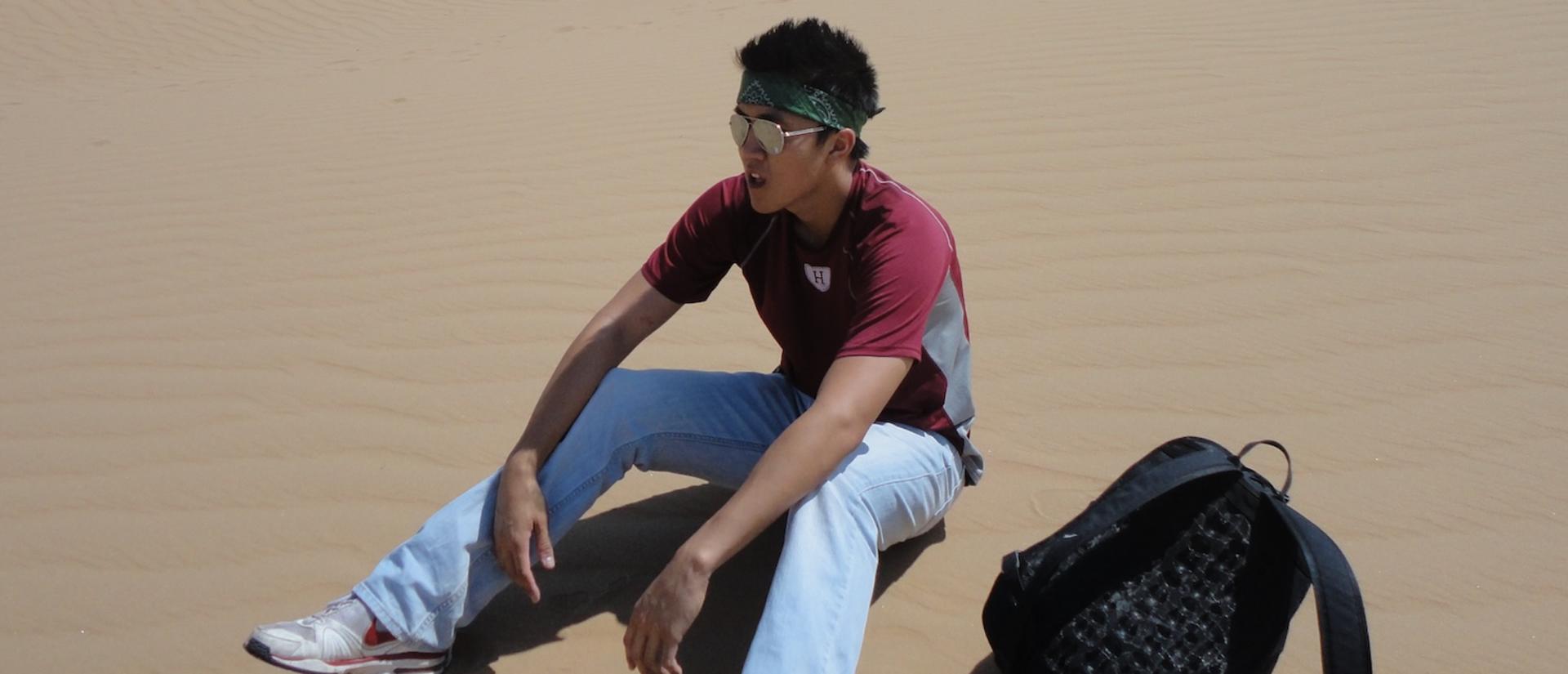 A person wearing sunglasses and a bandana sits on sand dunes with a backpack beside them under the bright sun.
