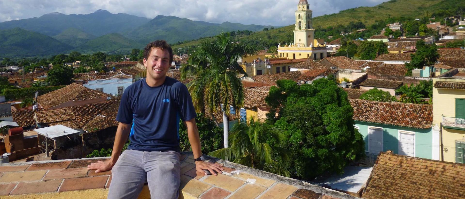 A person sits on a brick ledge smiling, with a town of tiled rooftops, palm trees, and mountains in the background.