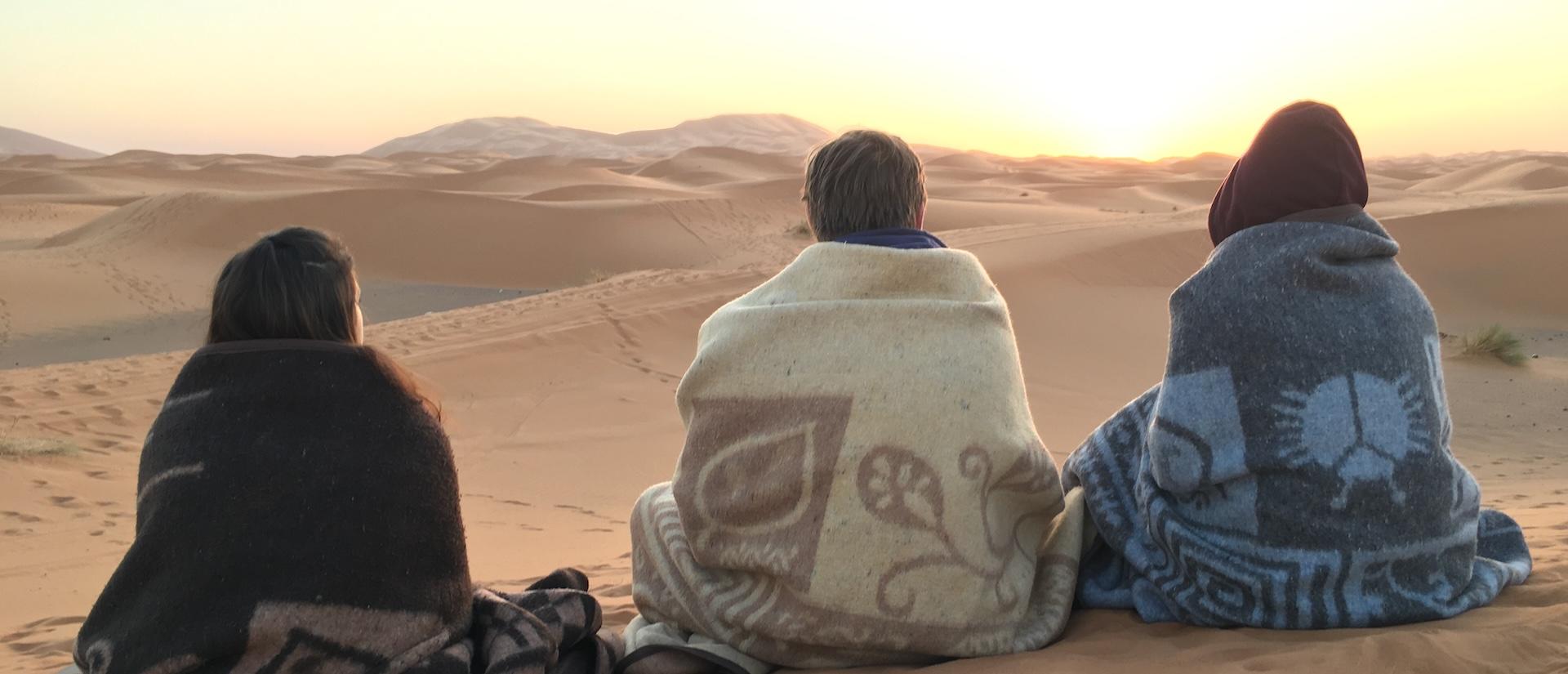 Three people sit wrapped in blankets on desert sand dunes, watching the sun set over the horizon.