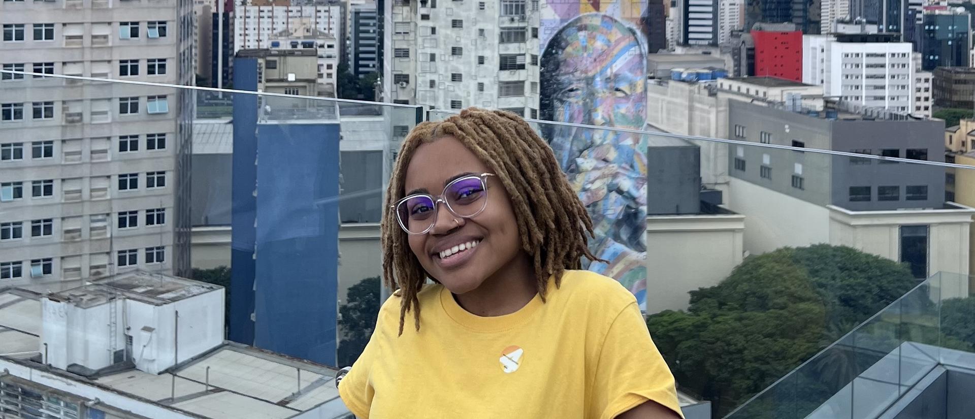 A smiling person in a yellow shirt poses on a rooftop with a city skyline and a colorful mural in the background.