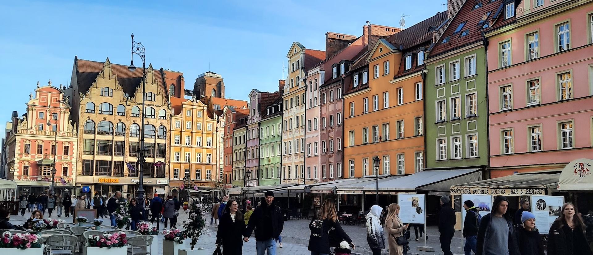 People walk through a lively square lined with colorful historic buildings and outdoor cafés.