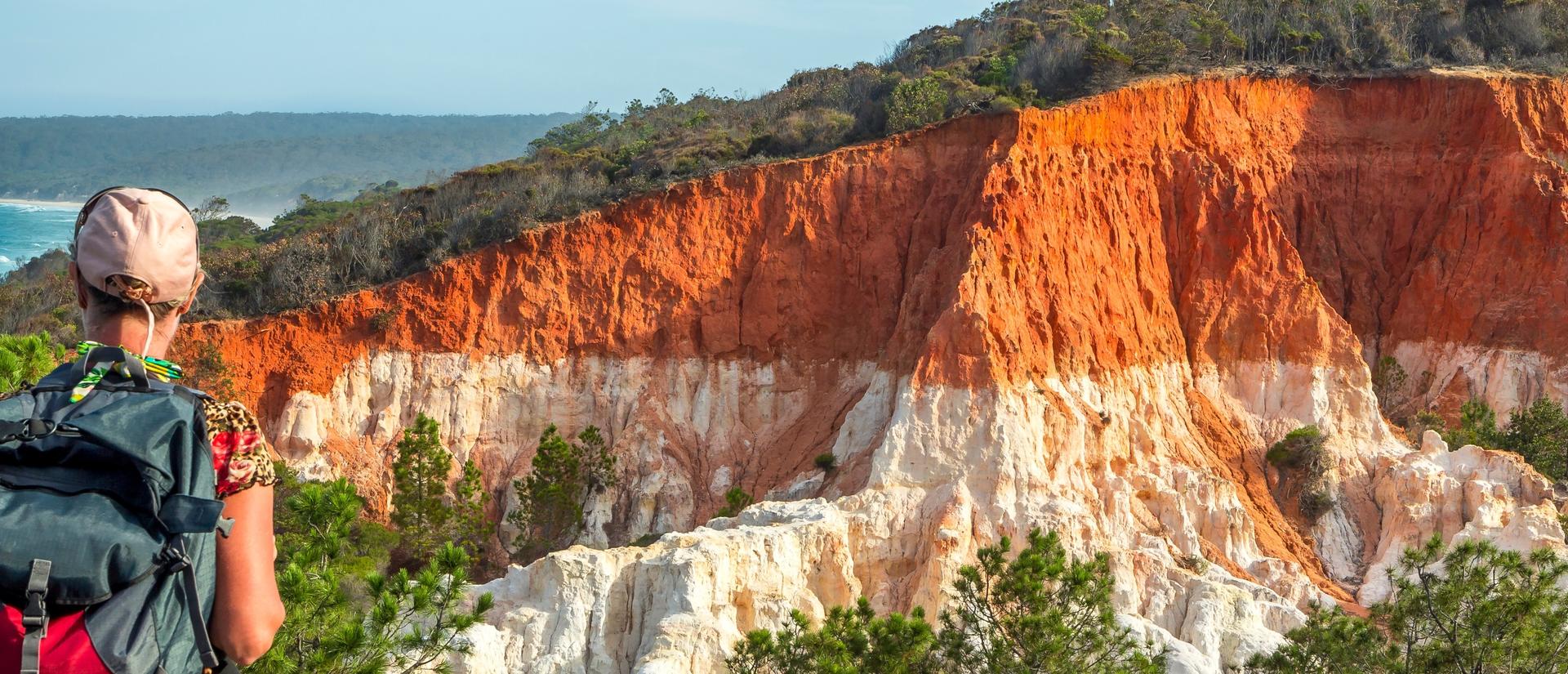 A hiker with a backpack looks out over colorful cliffs near the ocean.