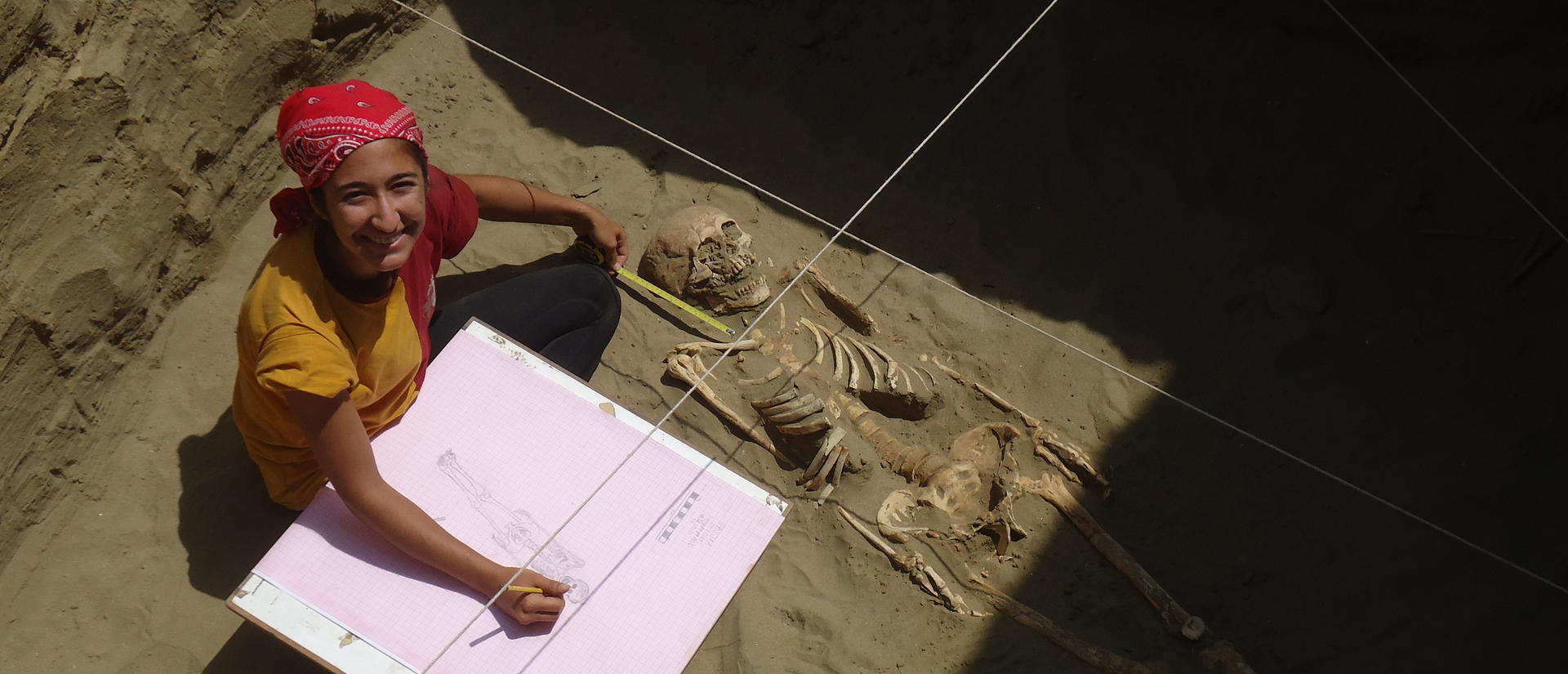 An archaeologist sketches and measures a skeleton at an excavation site.