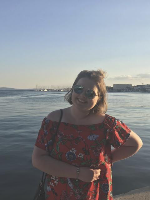 A smiling person in sunglasses and a red floral dress poses by the waterfront at sunset with boats and city buildings in the background.