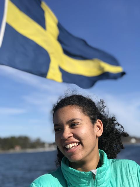 A smiling person in a teal jacket stands outdoors with the Swedish flag waving in the background.