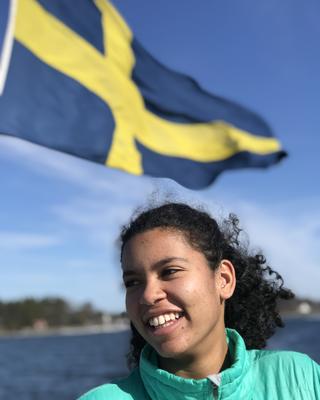 A smiling person in a teal jacket stands outdoors with the Swedish flag waving in the background.